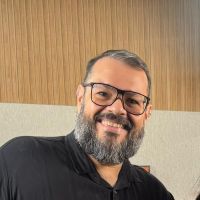 Foto de Wagner: homem de pele branca sorrindo para a câmera, com barba cheia e grisalha, usando óculos de armação escura e camisa preta, em ambiente interno com parede de madeira ao fundo.