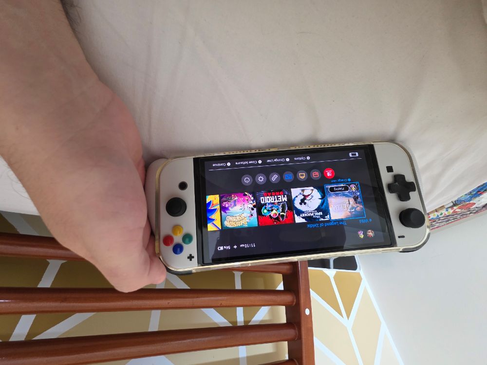 Pessoa segurando um console portátil Nintendo Switch com controles acoplados, exibindo a tela inicial com jogos; o dispositivo está sobre uma superfície clara, com parte de uma cama e uma estrutura de madeira ao fundo.