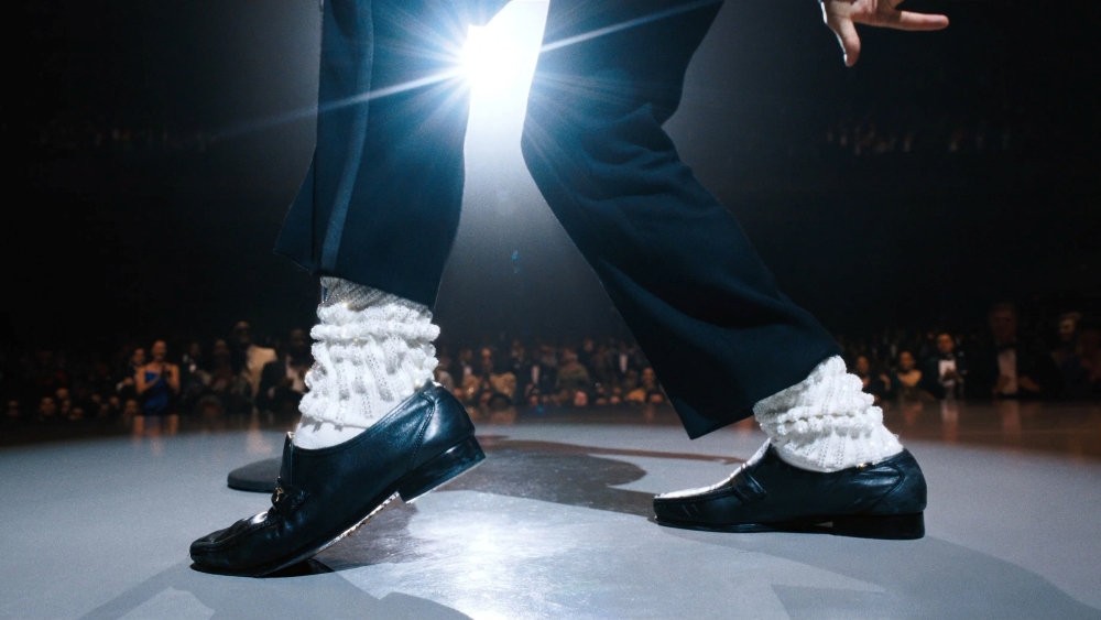 "Close nos pés de um dançarino no palco, em posição inclinada típica de um passo icônico. Ele usa sapatos sociais pretos bem polidos e meias brancas brilhantes, com textura franzida, contrastando fortemente com a roupa escura.
A perna da frente está esticada e apoiada no calcanhar, enquanto a de trás sustenta o peso, criando a ilusão de deslizamento. No fundo, desfocado, há uma plateia observando. Um feixe de luz intensa vem de trás, criando um efeito de halo e destacando o movimento e o brilho do figurino."
