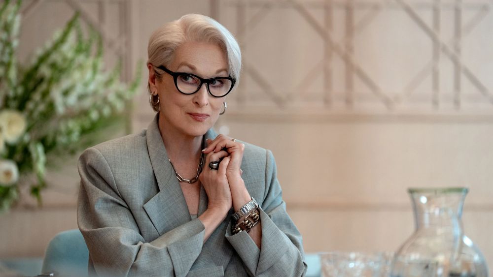 "Mulher de aparência elegante, com cabelo curto grisalho bem alinhado e usando óculos de armação escura, está sentada à mesa em um ambiente sofisticado. Ela veste um blazer claro e acessórios discretos, como pulseiras e anéis, e mantém as mãos próximas ao rosto, olhando de lado. Ao fundo, o espaço é claro e minimalista, com elementos decorativos suaves, como flores e um jarro de vidro."
