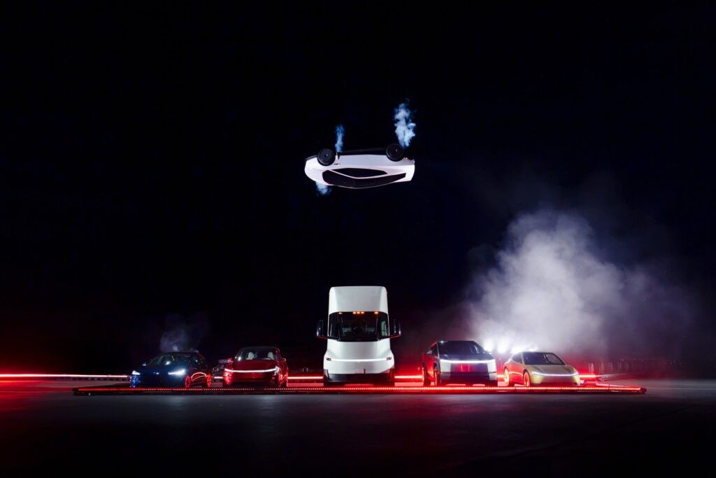 Roadster inverted in midair above Tesla vehicles and a Semi