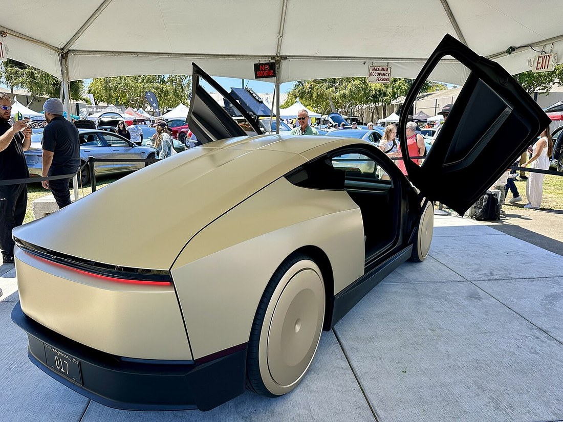 Gold Tesla Cybercab with a butterfly door open at an outdoor event, CYBERCAB vanity plate visible
