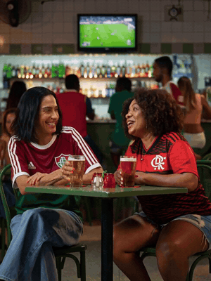 Two friends in Flamengo and Fluminense shirts laugh and clink beer glasses in a Rio bar