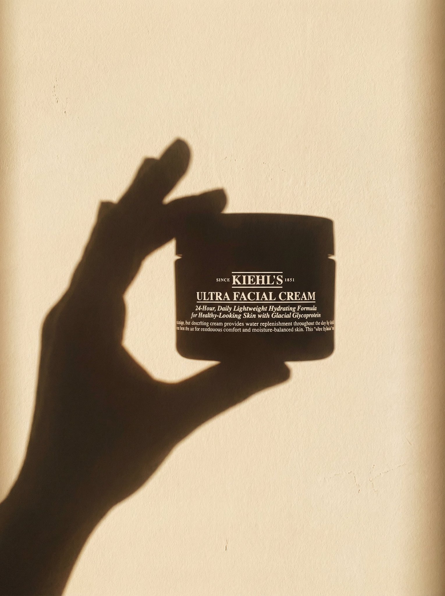 A minimal sunlit wall. Sharp, elongated shadow of a human hand holding the exact product from the uploaded image. Rec…